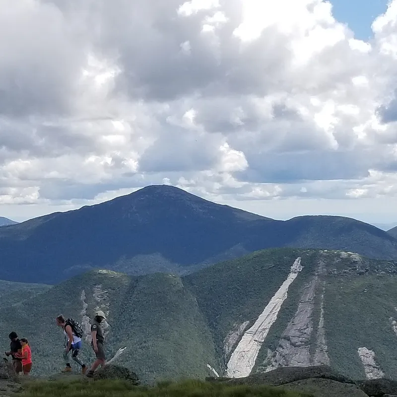 Hikers on the rocky summit of Algonquin Peak looking across to Mount Marcy and Mount Colden